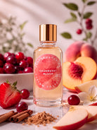 A clear glass perfume bottle with a gold cap and a pink circular label reading 'Camper Haven Custom Collection Cranberry Blush,' styled with cranberries, strawberries, peaches, cinnamon sticks, and sugar on a light surface.