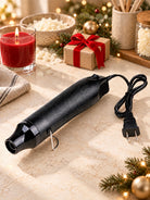 A black handheld heat gun for candle making with a textured grip and attached power cord, styled on a beige marble-like surface with holiday decorations including a red candle, gift box, pine branches, and lights in the background.
