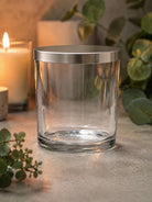 Clear glass Ilite candle vessel with metallic Ilite lid on a surface with candles and greenery in the background