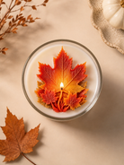 A glass jar candle with white wax and a decorative autumn leaf design in the center, featuring a large orange and yellow maple leaf with smaller red and orange leaves around it. The candle is styled on a beige surface with dried leaves and a small pumpkin in the corners.
