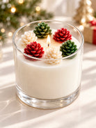 A lit white candle in a clear glass jar, decorated with five mini pinecones in red, green, and cream, set against a softly blurred holiday background with warm lights.