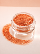 Shimmering orange diamond sparkle dust in a small clear round jar, with some of the diamond sparkle dust spilled in a curved line beside it on a soft light beige background.