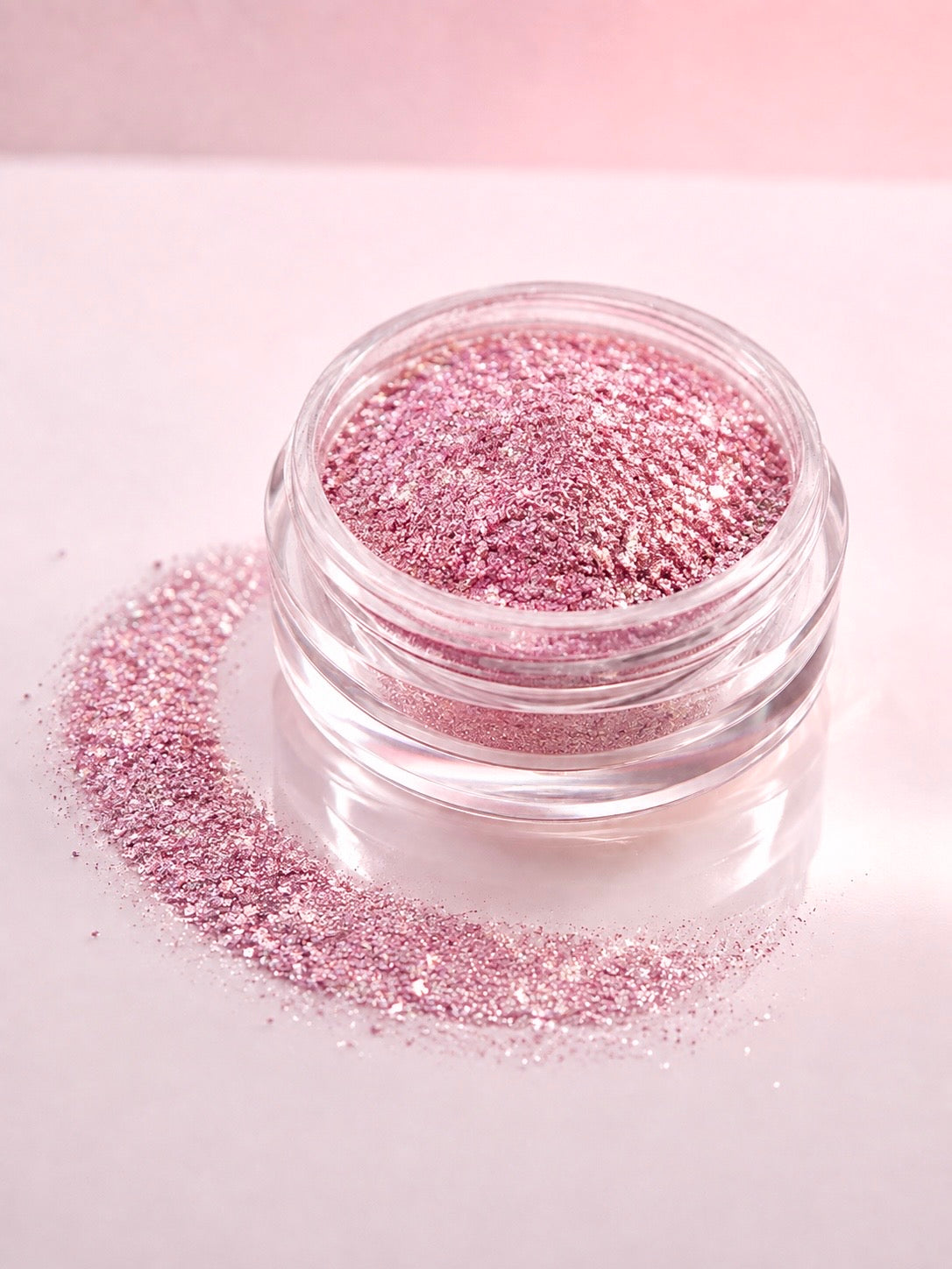 A small clear jar filled with sparkling pink diamond sparkle dust, with loose pink diamond sparkle dust scattered around it on a pale pink background.