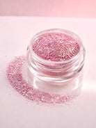 A small clear jar filled with sparkling pink diamond sparkle dust, with loose pink diamond sparkle dust scattered around it on a pale pink background.
