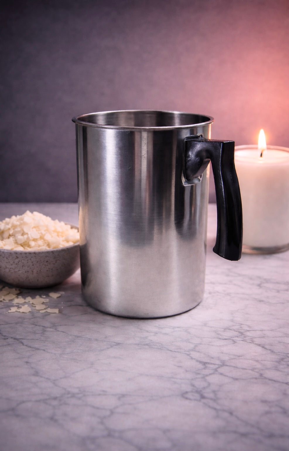 Metal pouring pitcher for candle maker with a black handle on a marble surface next to a bowl of rice and a lit candle.