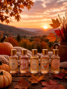 Five bottles of pumpkin style fragrance oils for candle making on a wooden surface with pumpkins and autumn leaves, set against a scenic fall sunset.