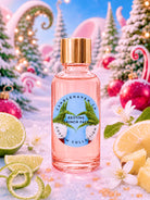 A pink glass bottle of fragrance oil for candle making with a gold cap and a circular blue-and-green label reading 'CandleHaven Resting Grinch Face' and 'Custom Collection,' styled in a snowy holiday scene with citrus slices, ornaments, candy canes, and lights.