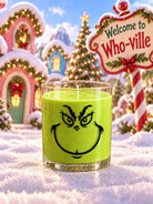A green Grinch-themed candle in a clear glass jar sits in a snowy, festive village scene with a 'Welcome to Who-ville' sign and Christmas houses in the background.