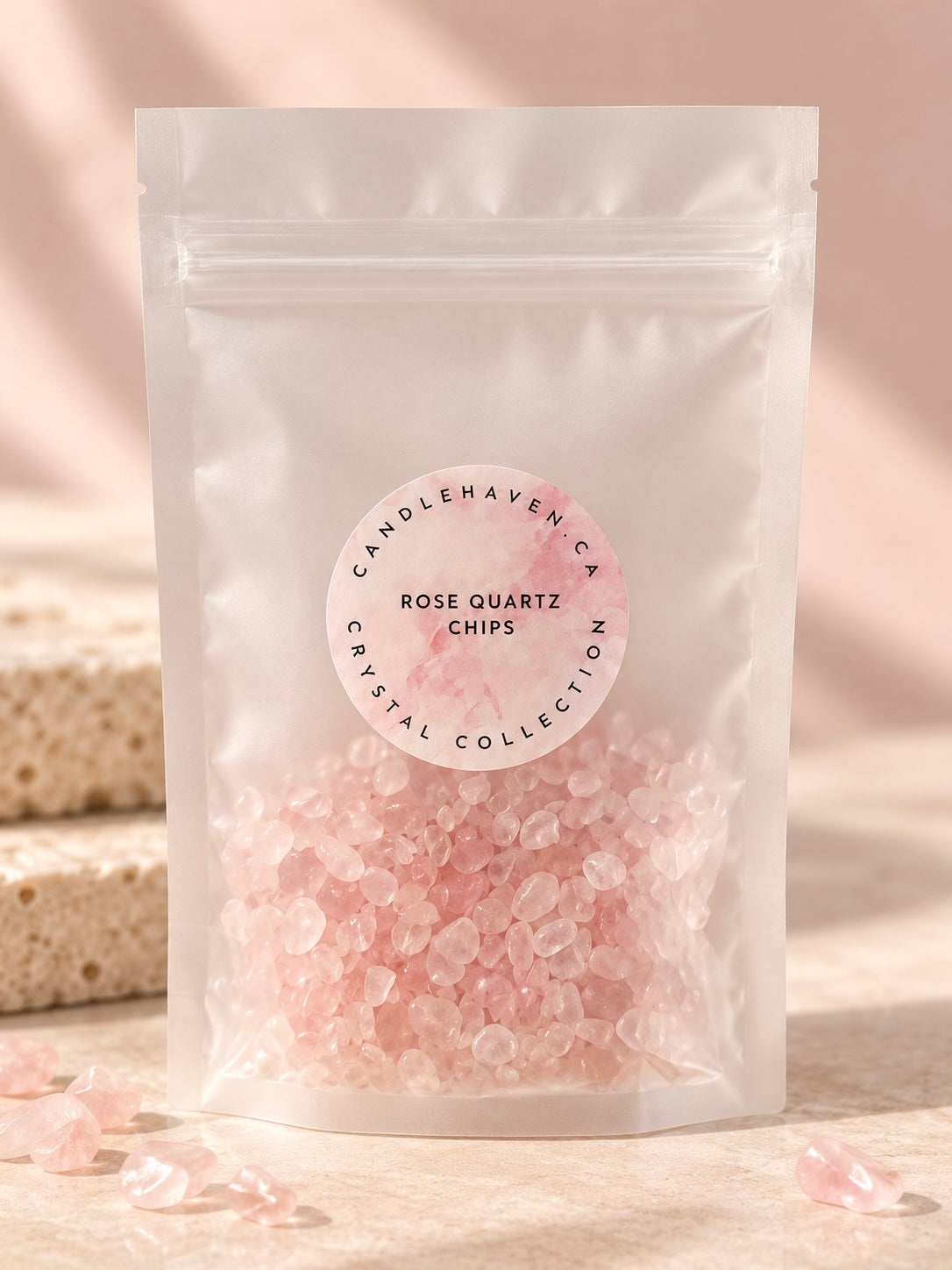 A clear stand-up pouch containing pink rose quartz chips, with a round white label reading 'Candlehaven CA Rose Quartz Chips Crystal Collection.' The pouch is photographed on a warm beige surface with soft shadows and a few loose pink crystal pieces around it.