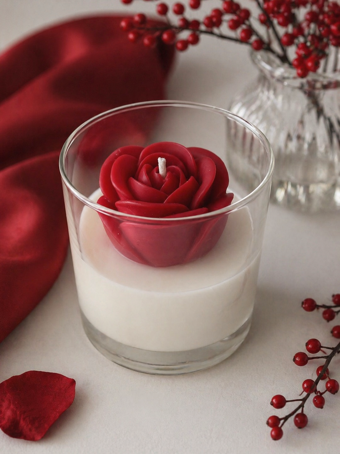 White candle with a red rose-shaped candle inside, surrounded by red berries and a red fabric background.