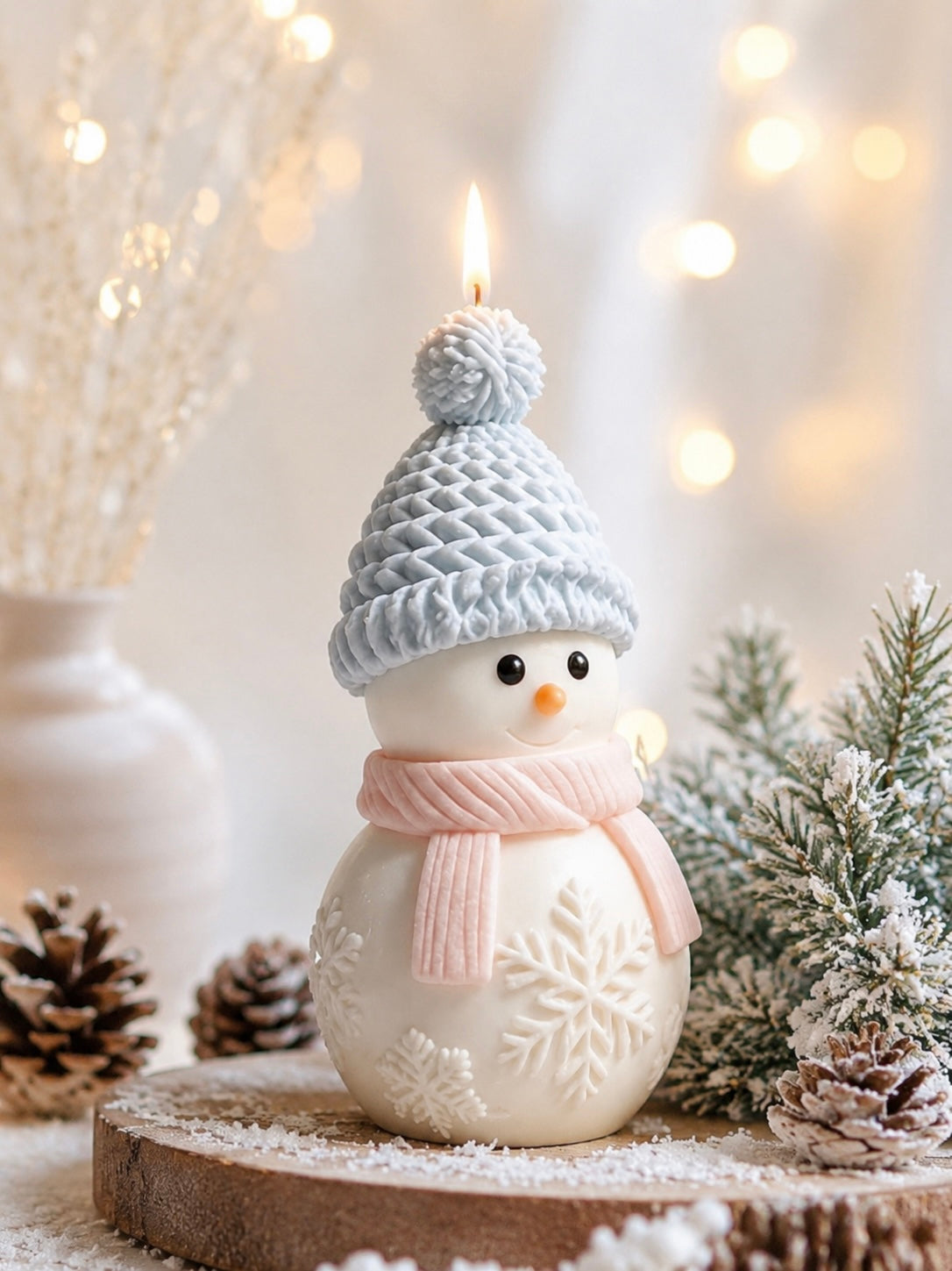 A snowman-shaped candle with a light blue knit hat and pink scarf, displayed on a rustic wooden slice with pinecones and frosted greenery against a warm, softly blurred holiday background.