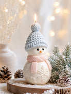 A snowman-shaped candle with a light blue knit hat and pink scarf, displayed on a rustic wooden slice with pinecones and frosted greenery against a warm, softly blurred holiday background.