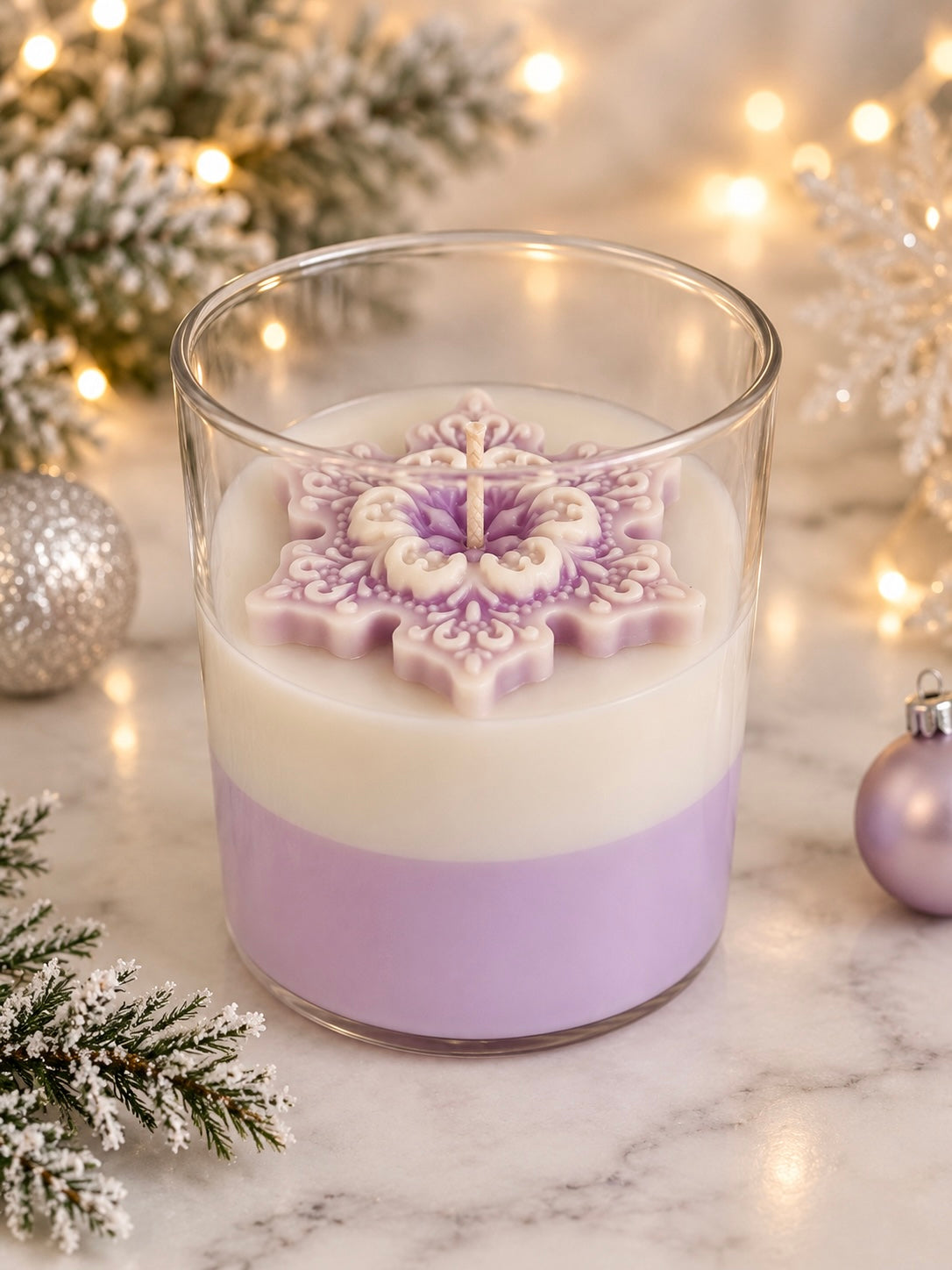 A layered candle in a clear glass jar sits on a marble surface with holiday decorations, including a white snowflake-shaped wax top, a lavender lower layer, and warm string lights in the background.