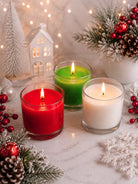 Three lit candles in clear glass jars, one red, one green, and one white, depicting wax dye chips for colouring candles arranged on a marble surface with Christmas decorations including pine branches, red berries, a small white tree, and a house-shaped ornament.