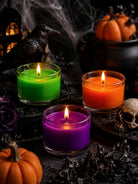 Three lit glass candles in green, orange, and purple depicting wax dye chips for colouring candles are arranged on a dark surface with Halloween props including pumpkins, a skull, dried leaves, a black cauldron, a raven, and cobwebs.