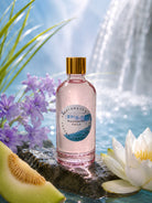 Bottle of 'Whispering Falls' fragrance oil by candlehaven with a waterlily and melon on a reflective surface.