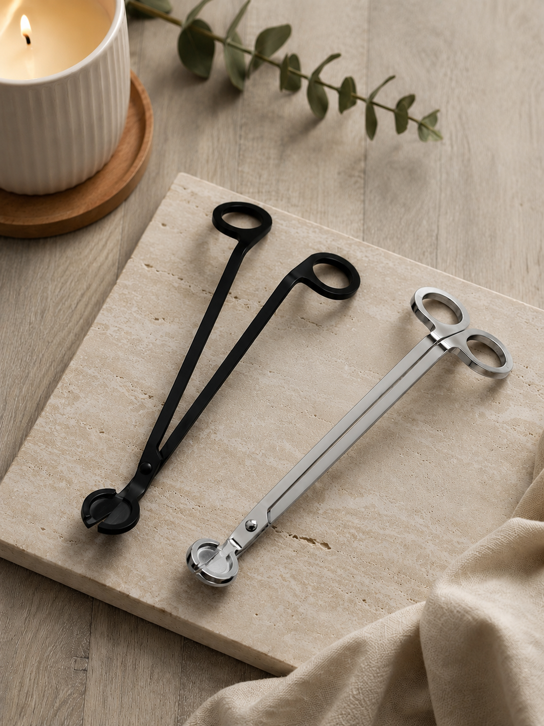 One black and one silver pair of wick trimmers on a wooden surface with a candle and leaves in the background.