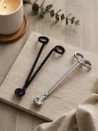 One black and one silver pair of wick trimmers on a wooden surface with a candle and leaves in the background.