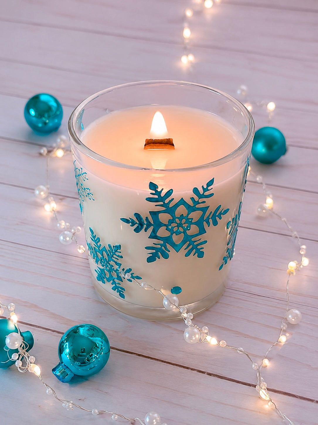 Candle with blue snowflake design on a wooden surface with decorative lights and ornaments.