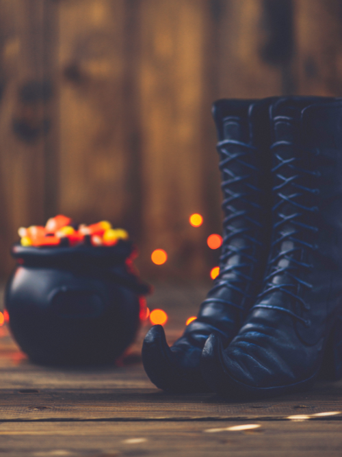 Black witch boots and a cauldron with colorful candies on a wooden surface with blurred lights in the background depicting candle fragrance oil Witch, Please!