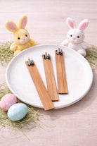 Three wooden wicks for candle making on a white plate with Easter-themed decorations including stuffed bunnies and eggs.