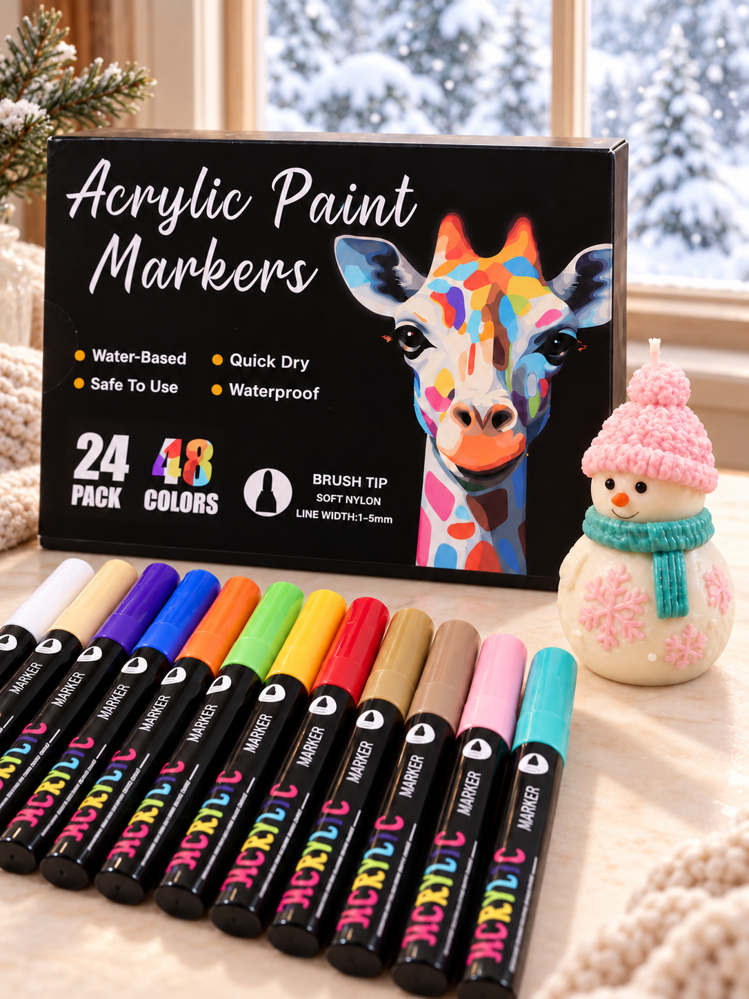 Acrylic paint marker box and colourful markers beside a snowman candle with pink details and a teal scarf, near a snowy window.