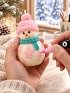 Hand holding a snowman candle while a pink acrylic marker colours a raised snowflake, with a pink hat and teal scarf.