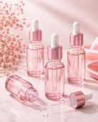 Three pink glass dropper bottles with metallic rose gold caps and white droppers, arranged on a light marble surface with soft pink floral decor in the background.